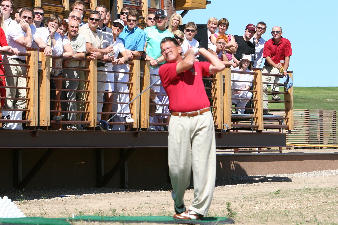 Brett Brasier teeing off at a driving range with a crowd watching from a clubhouse deck at Capitals Golf Club, Lithuania